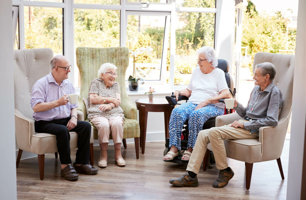 A group of seniors sitting in a semicircle in a common area, drinking tea or coffee and laughing