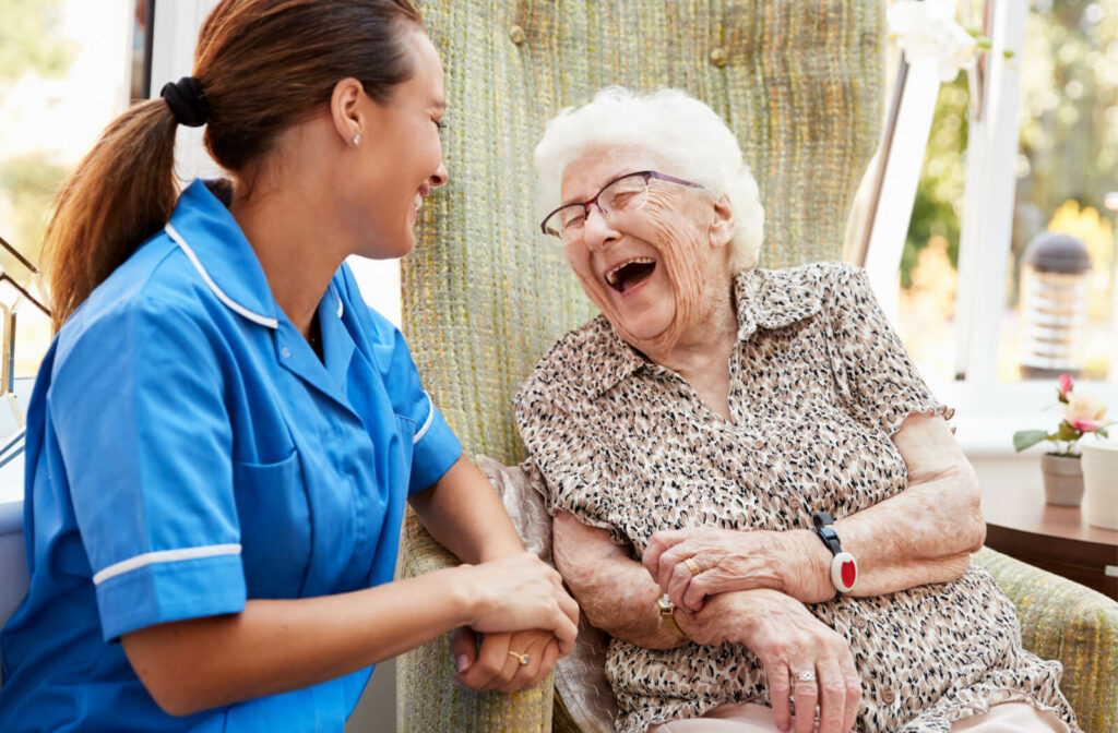 A senior woman in a senior living facility sitting on a chair smiling and having a conversation with a nurse.