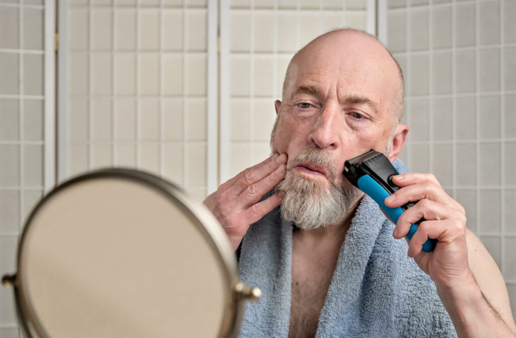 An older adult man shaving his beard in front of a small mirror using an electric razor.