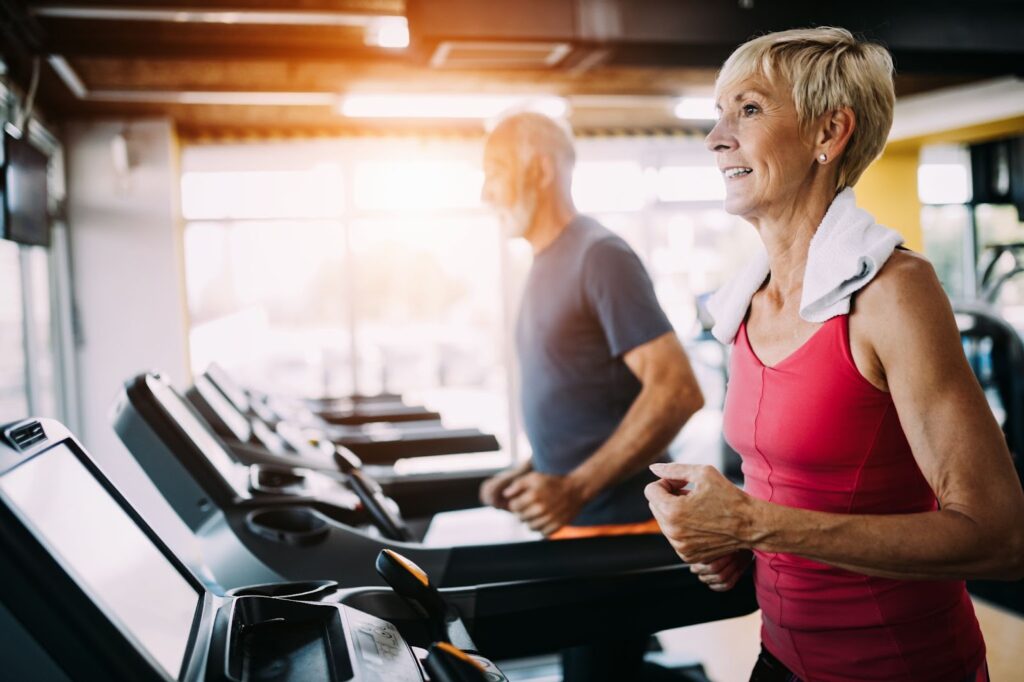 A senior couple running beside each other on treadmills in a brightly-lit exercise room.