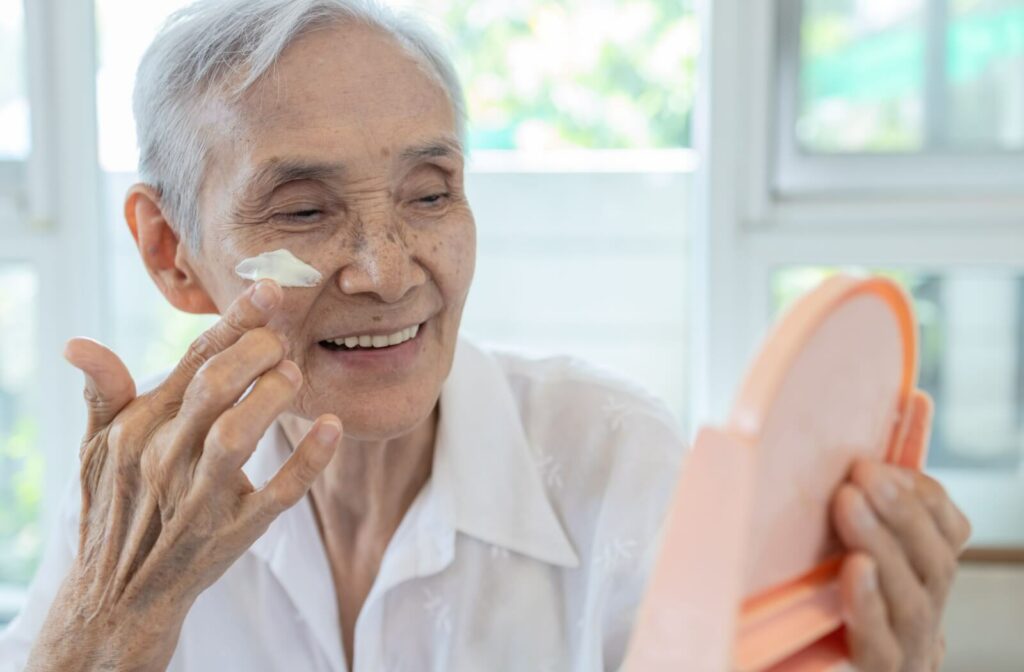 An older woman holds a circular mirror to look at her reflection while applying moisturizer to her cheeks