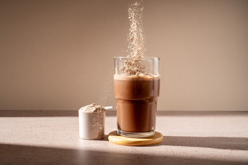  A chocolate meal replacement shake is on a coaster beside a full scoop of protein powder.